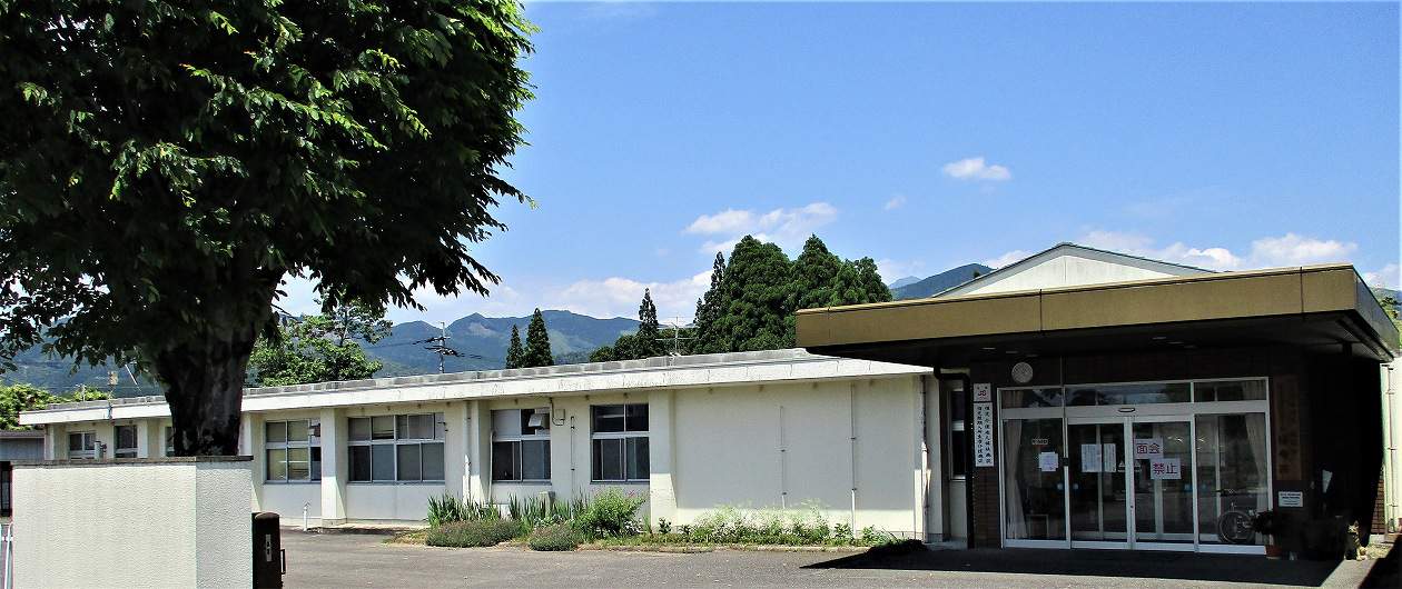 平屋建ての介護施設の外観。白壁で統一され、駐車場側から見た建物全景。山々を背景に。